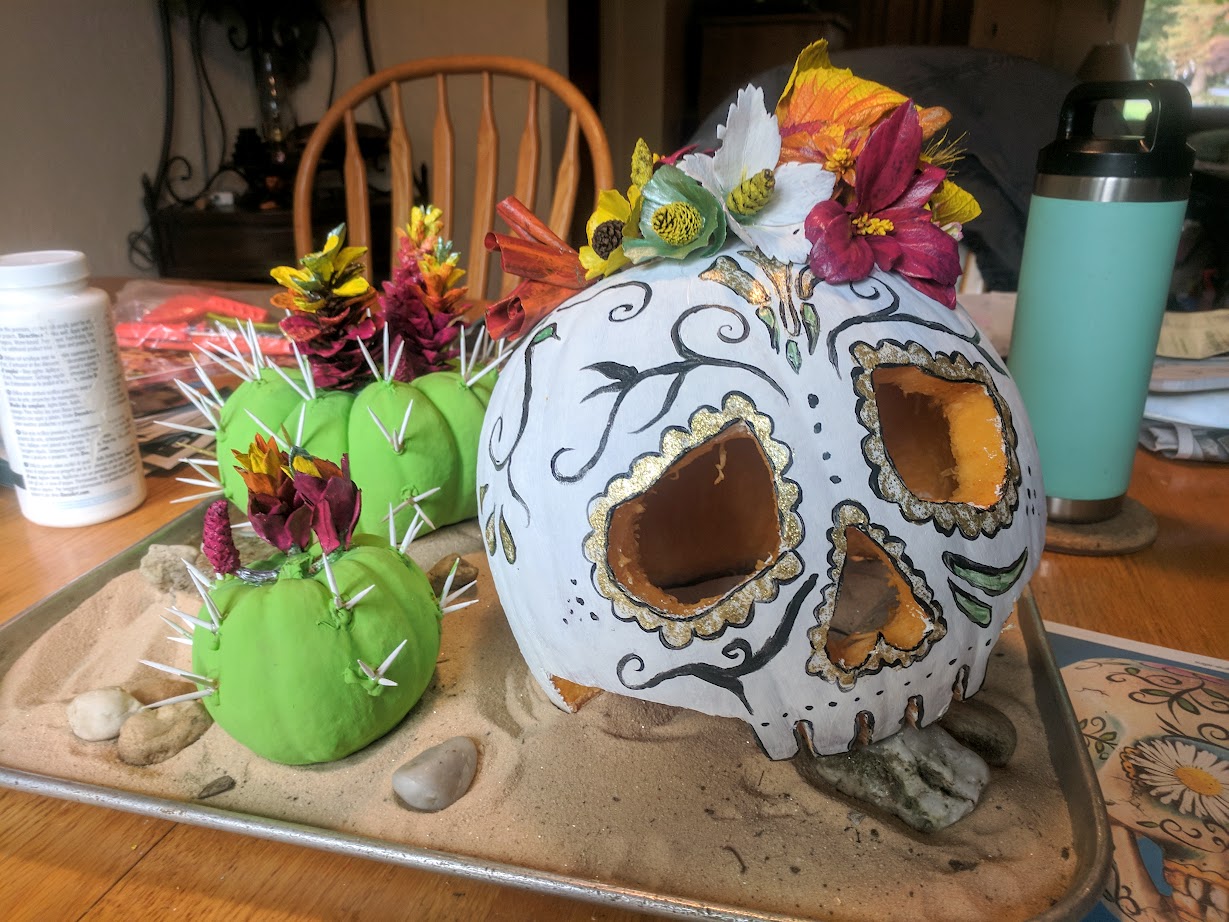 a pumpkin carved and painted to look like a skull, decorated with colorful flowers made from leaves, surrounded by blooming cacti made from squatty pumpkins