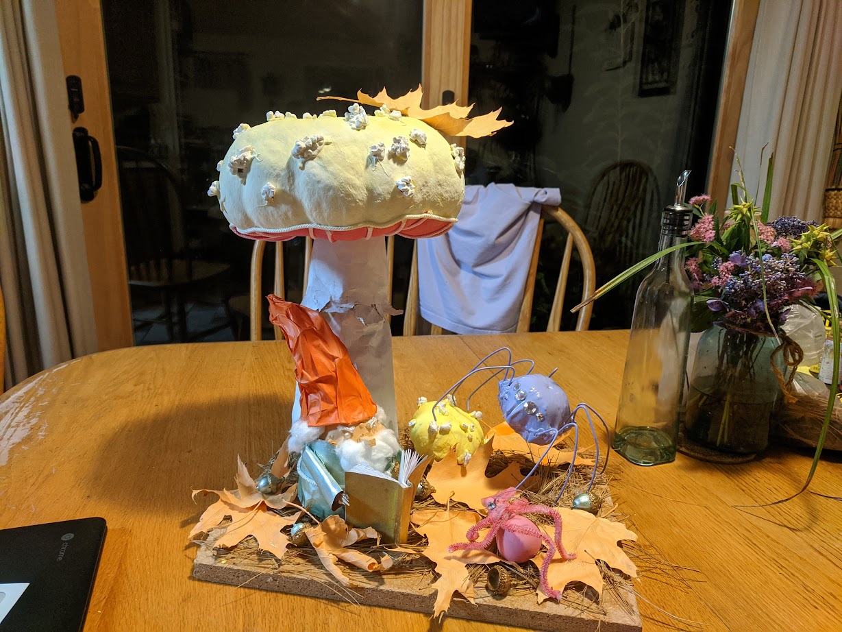a gourd decorated to look like a gnome reads a story to some gourd bugs under a pumpkin decorated to look like a mushroom.