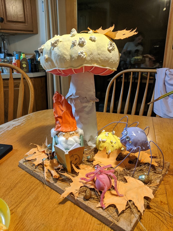 a gourd decorated to look like a gnome reads a story to some gourd bugs under a pumpkin decorated to look like a mushroom.